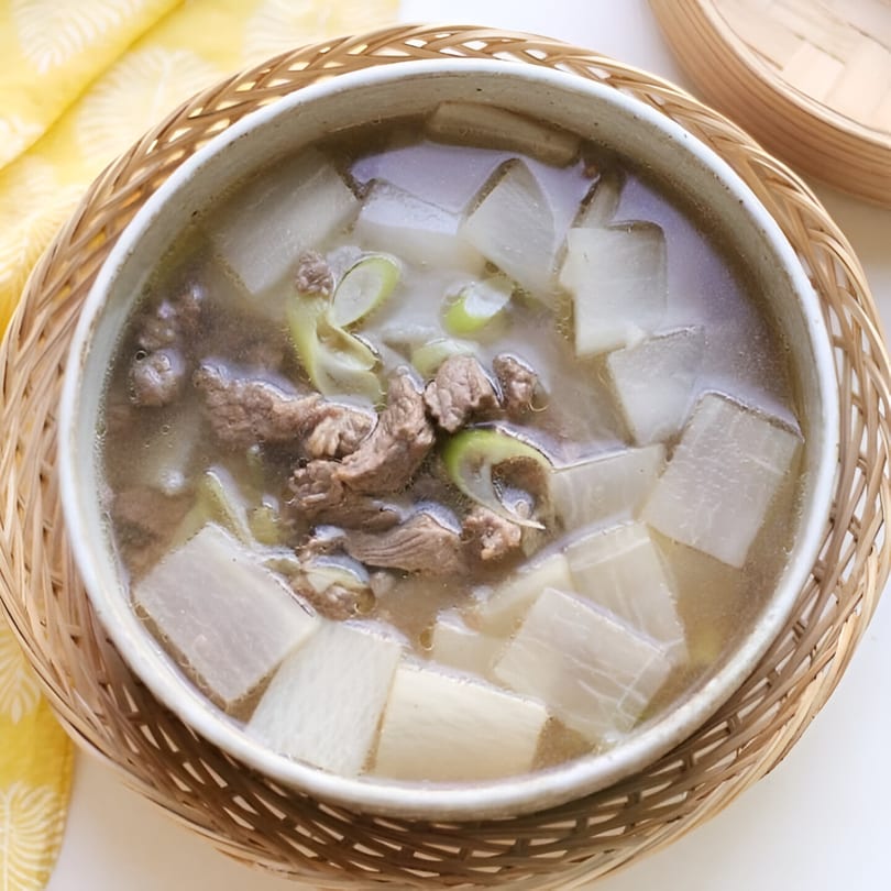A Korean beef and radish soup, known as Muguk, in a white bowl on a woven placemat.