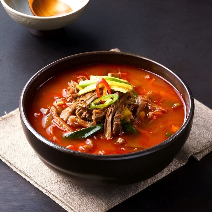 Spicy Korean Yukgaejang soup with shredded beef, vegetables, and chili peppers in a black bowl.