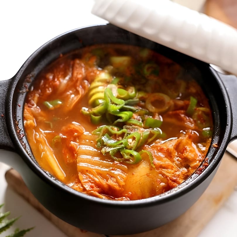 A black pot of Korean kimchi stew, garnished with sliced green onions and peppers.