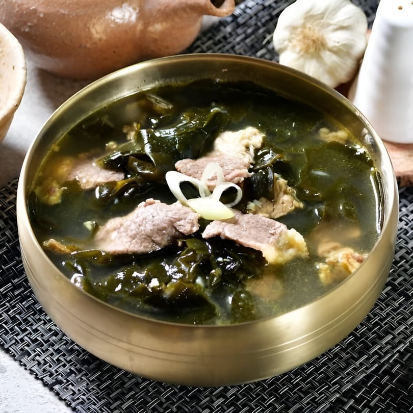 A brass bowl of Korean seaweed soup with beef and sliced onions, on a dark woven placemat.